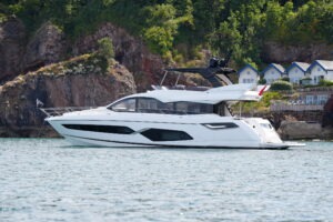 2021 Sunseeker Manhattan 68 yacht cruising near rocky coastline and houses.